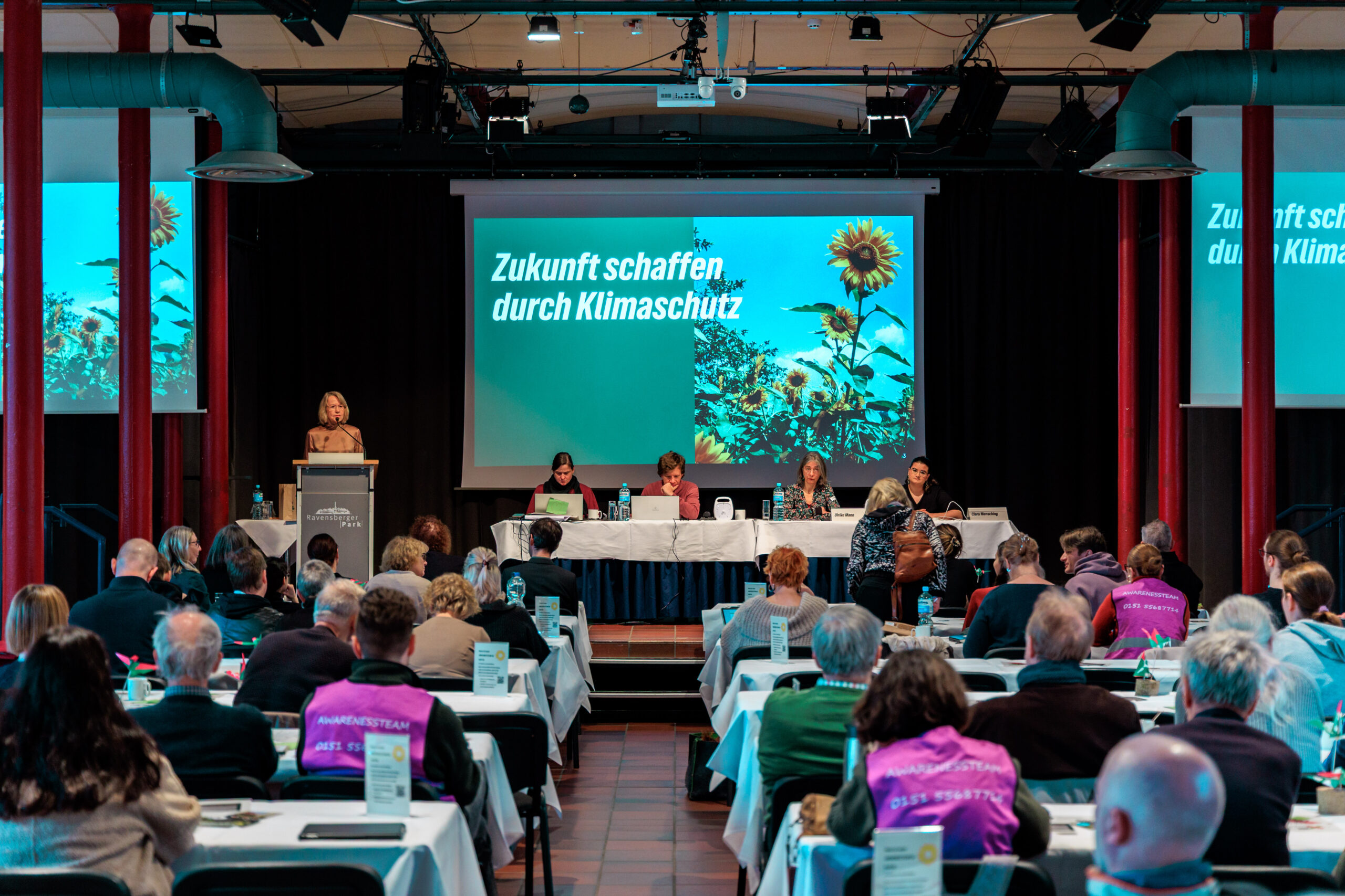 Zu sehen ist ein großer Saal mit Grünen Mitgliedern. Auf der Bühne spricht Brigitte Meier.